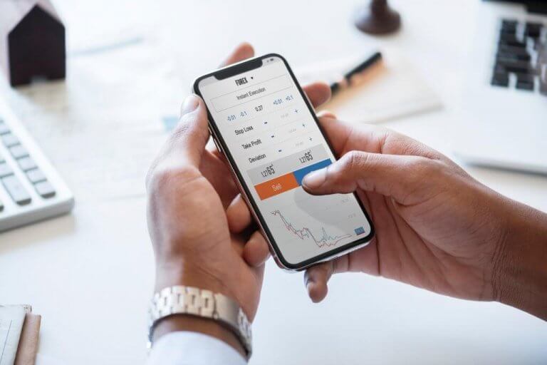 يدان لرجل يرتدي ساعة يد فضية اللون فوق الطاولة يحمل هاتف ذكي على شاشته تظهر واجهة منصة تداول بيضاء وأمامه أشياء أخرى